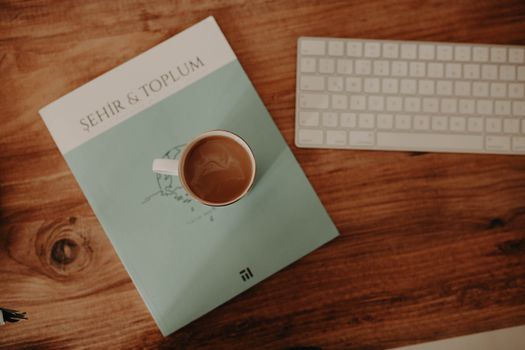 Flat lay of a coffee cup on a magazine with a keyboard nearby on a wooden table.