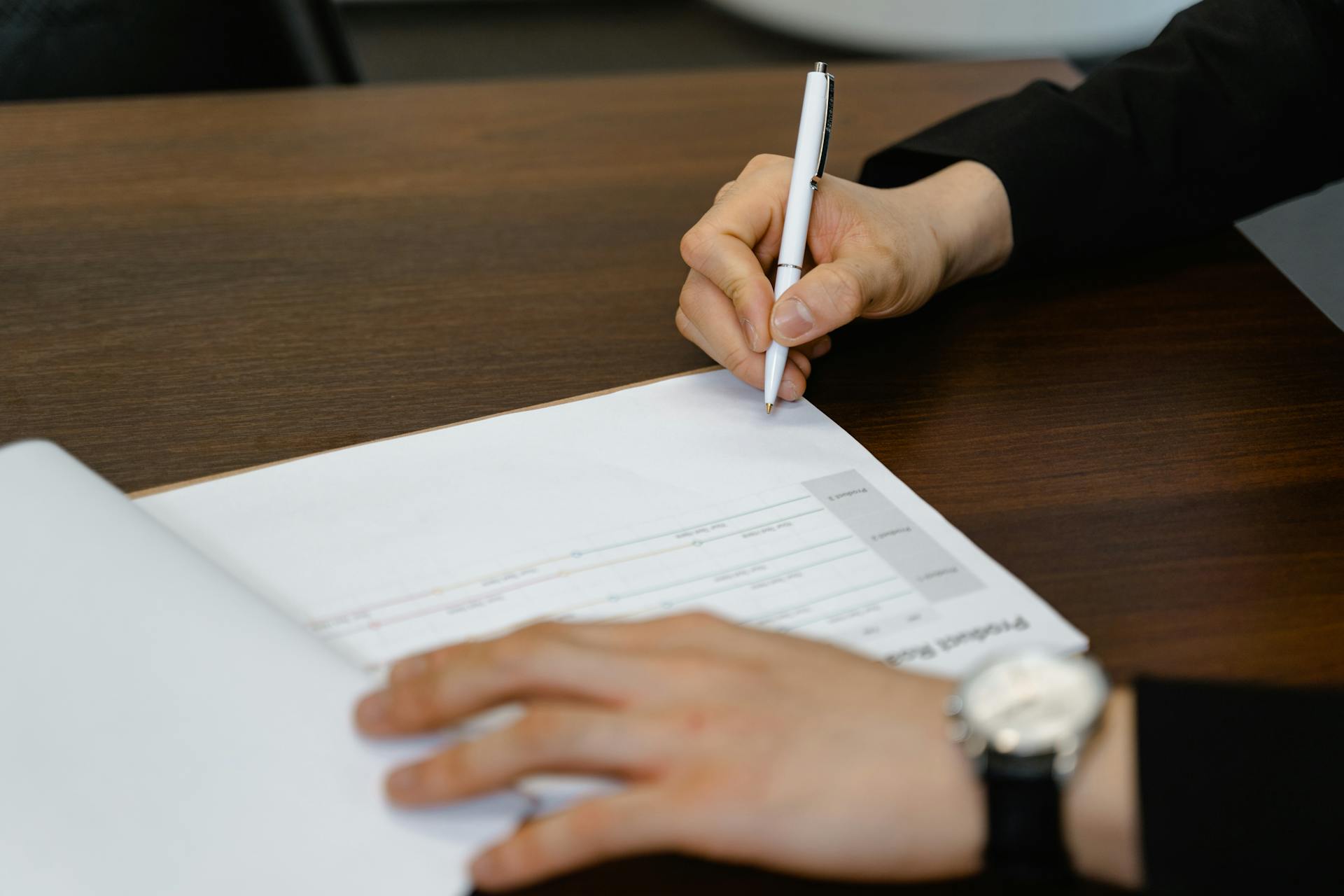 Businessperson writing on a document at a wooden desk, with focus on hands and pen.