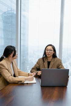 Two Asian businesswomen discussing work at a laptop in a modern office setting.