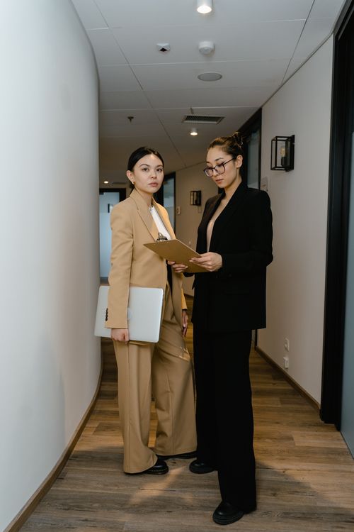 Free Two businesswomen in formal attire stand in an office hallway discussing work. Stock Photo