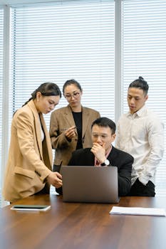 A group of colleagues engaged in a meeting, discussing around a laptop in a modern office.