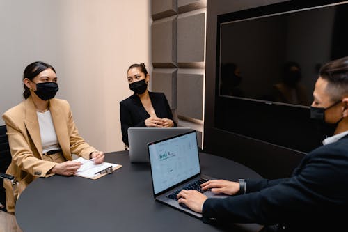 Two Women in Face Masks Sitting in Front of Man in Suit with Laptop
