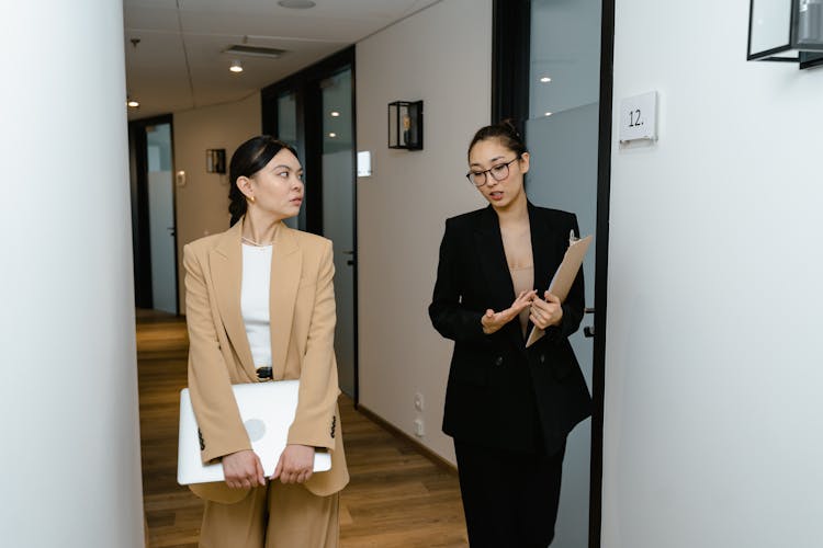 Woman In Glasses Talking To Woman With Laptop In Hallway