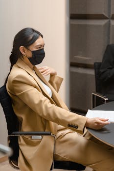Asian woman in formal attire wearing a mask during an indoor business meeting.