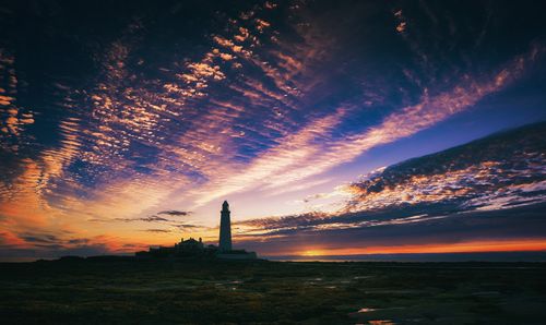 Free Silhouette of a lighthouse against a vibrant sunset and dramatic sky with scattered clouds. Stock Photo