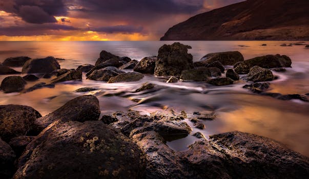 A breathtaking view of the ocean at sunset with rocks in the foreground and dramatic clouds.
