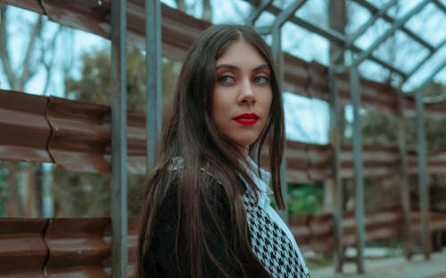 Side view of confident young female with long straight hair and bright red lips against metal construction looking away
