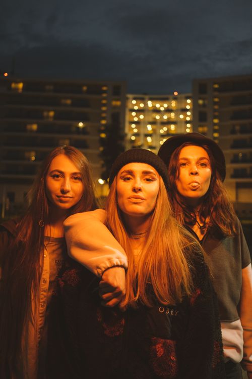 Free Three young women posing confidently at night against a backdrop of lit apartment buildings. Stock Photo
