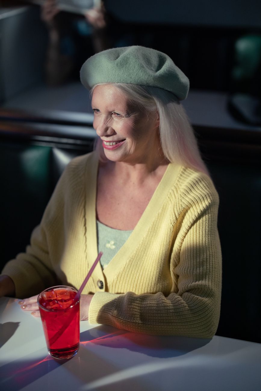 A cheerful senior woman in a beret and cardigan, enjoying a drink at a diner table.