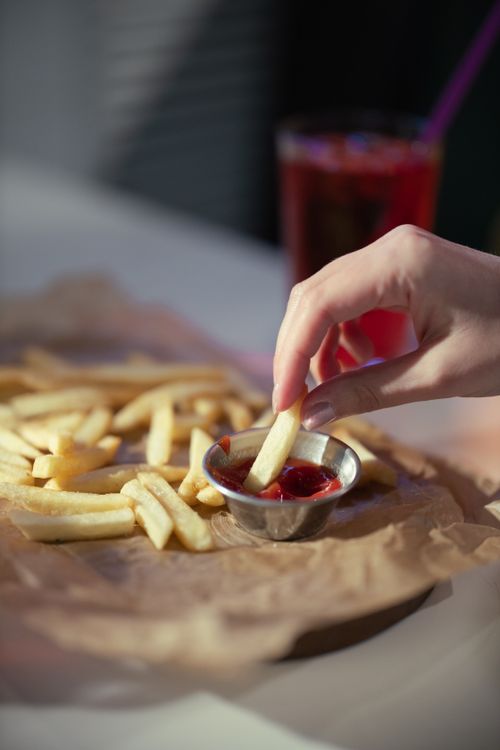 Free A close-up of a hand dipping french fries into a small bowl of ketchup, on a table outdoors. Stock Photo
