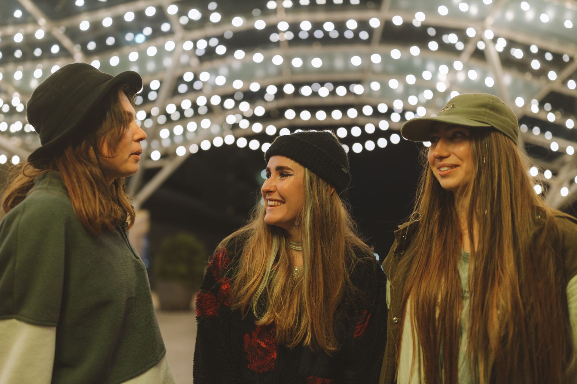 Three young women enjoying each other's company under twinkling canopy lights at night.