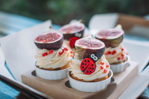 Tasty cupcakes with white frosting decorated with fresh cut figs and sprinkles placed in package on table against blurred background