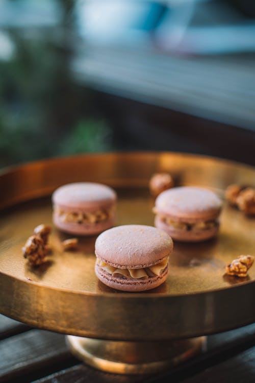 Delicious pink macarons with sweet filling served on round golden tray with decorations on wooden table on blurred background