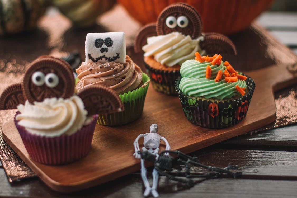 Tasty spooky Halloween cupcakes with sweet frosting decorated with cookies and marshmallow placed on cutting board on wooden table in light room