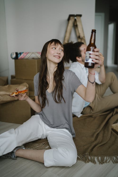 A Woman Sitting on Floor Holding Pizza and a Bottle of Beer