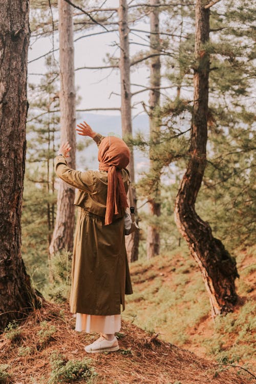 Back view full body of unrecognizable female in headscarf and trench coat standing among trees in woods