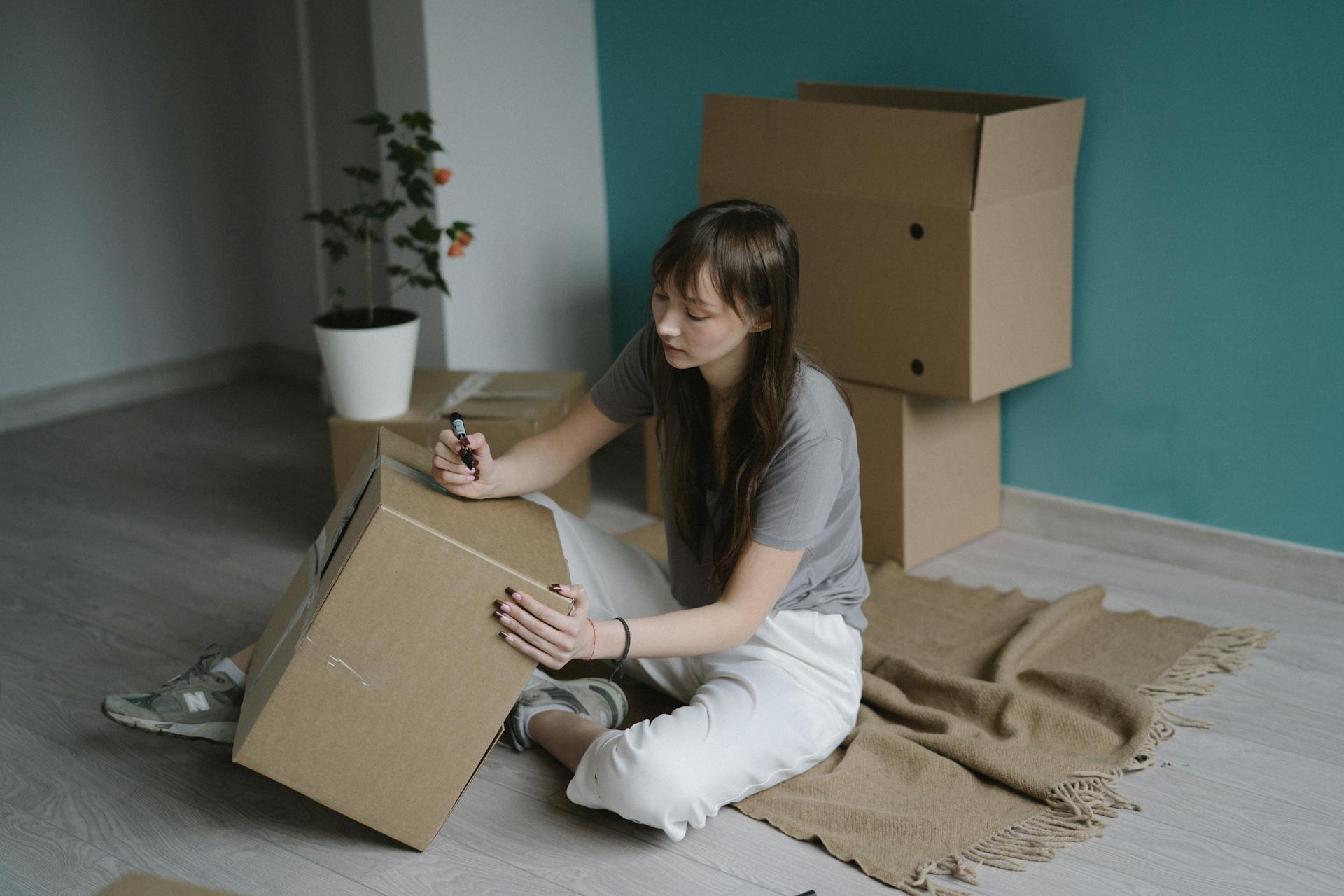 Young woman labeling a cardboard box while sitting on the floor. Moving or packing concept.