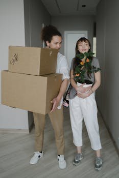 A young couple carrying boxes and a plant into their new home, symbolizing a fresh start.