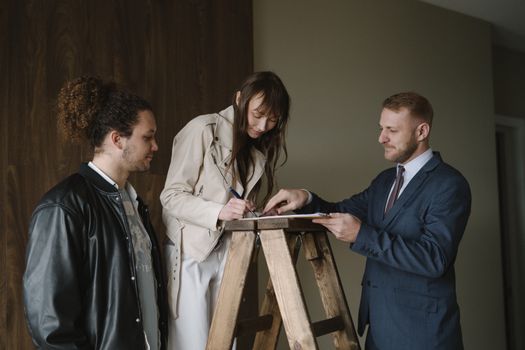 Three individuals signing a contract on a ladder inside a new house, symbolizing a real estate deal.