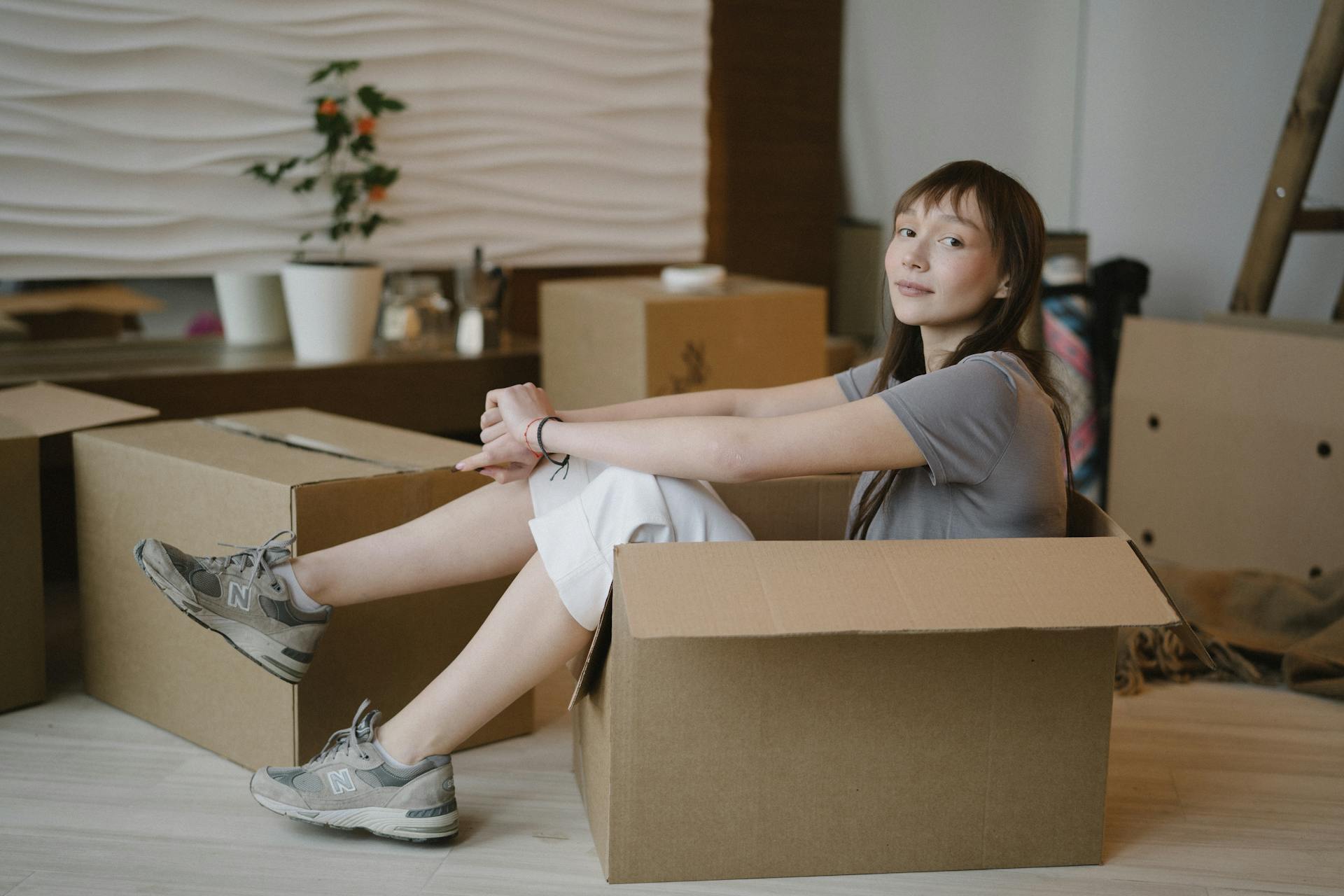 Woman sits in a cardboard box surrounded by moving boxes, symbolizing a new home journey.