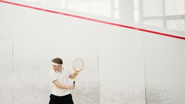 Active squash player in white sportswear preparing for a serve on indoor court.