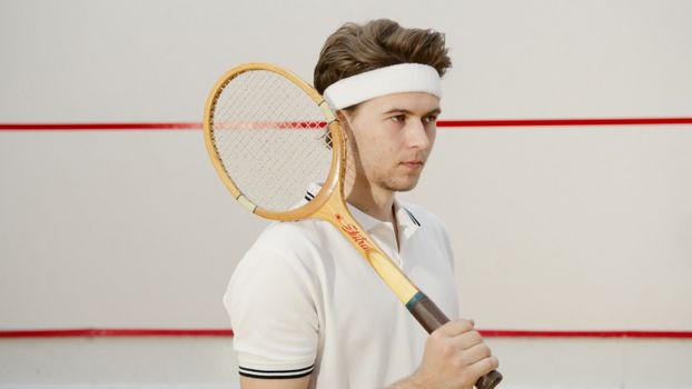 A young man in white polo and headband holding a vintage tennis racket indoors.