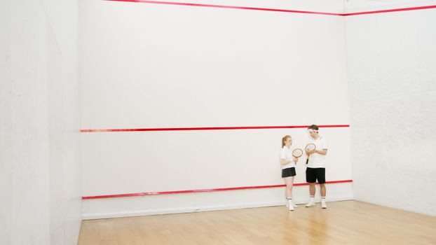 A young couple with rackets stands in an empty squash court, ready to play.