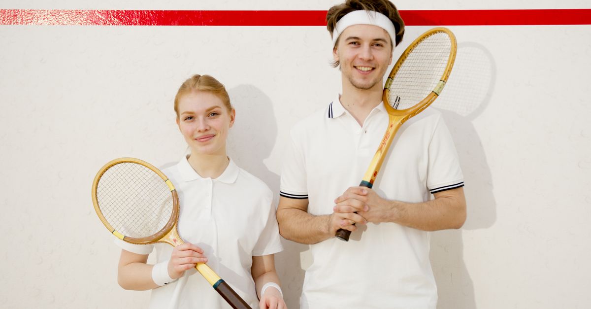 A young man and woman holding squash rackets and smiling indoors on a squash court.