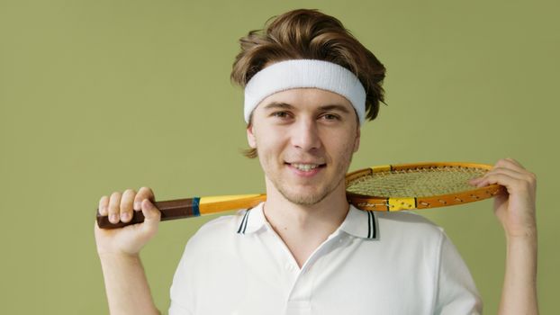 Smiling young man in a polo shirt posing with a vintage squash racket against a green background.