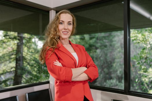Businesswoman in red blazer smiling confidently in office with greenery backdrop.