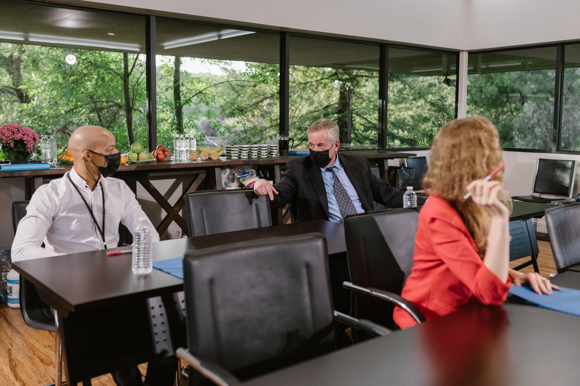 Business professionals wearing masks discuss in a sunlit conference room with spaced seating.