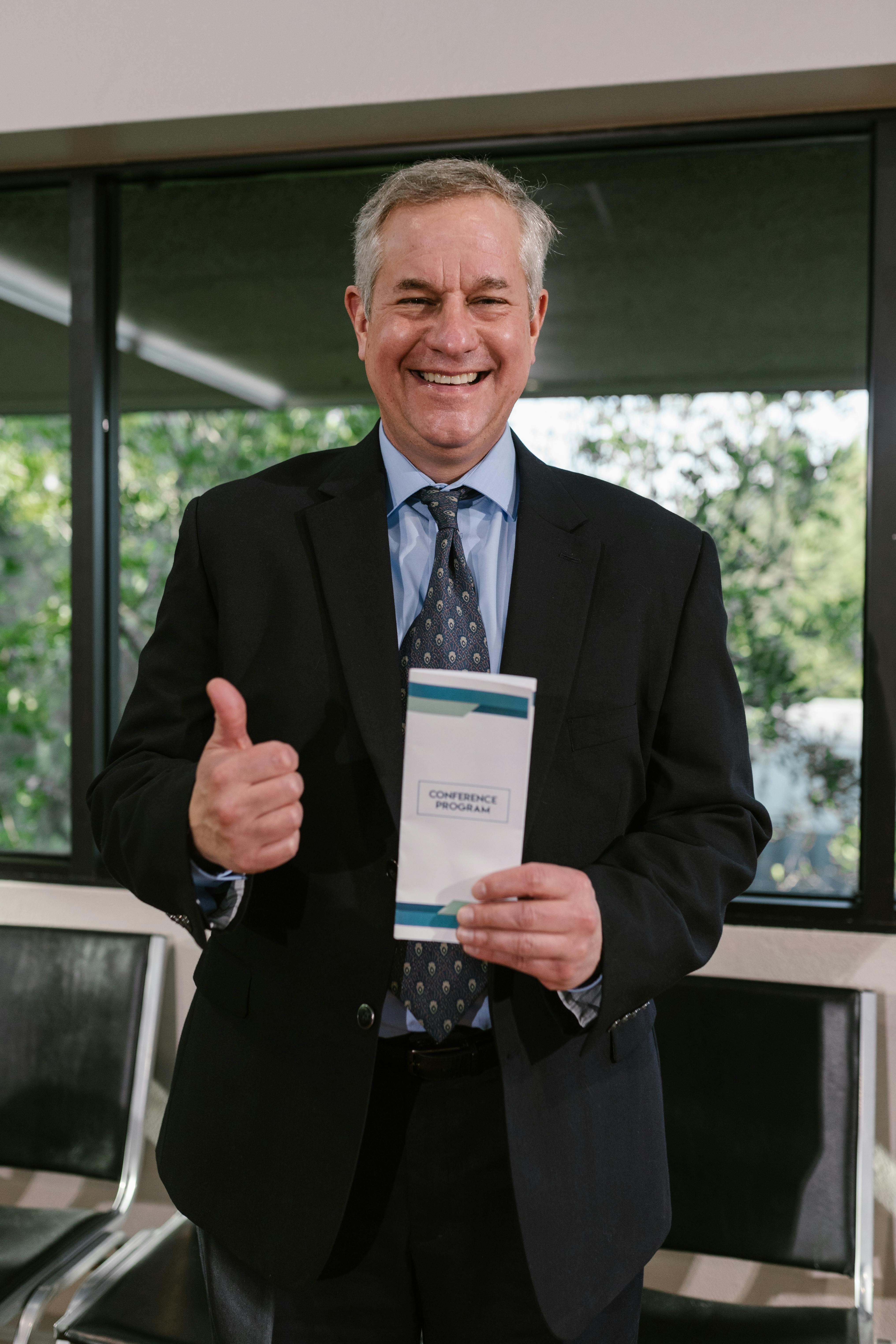 Smiling businessman in suit holding a conference brochure, giving thumbs up indoors.
