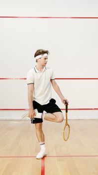 A man performs a pre-play stretch on a squash court, holding a vintage racket.