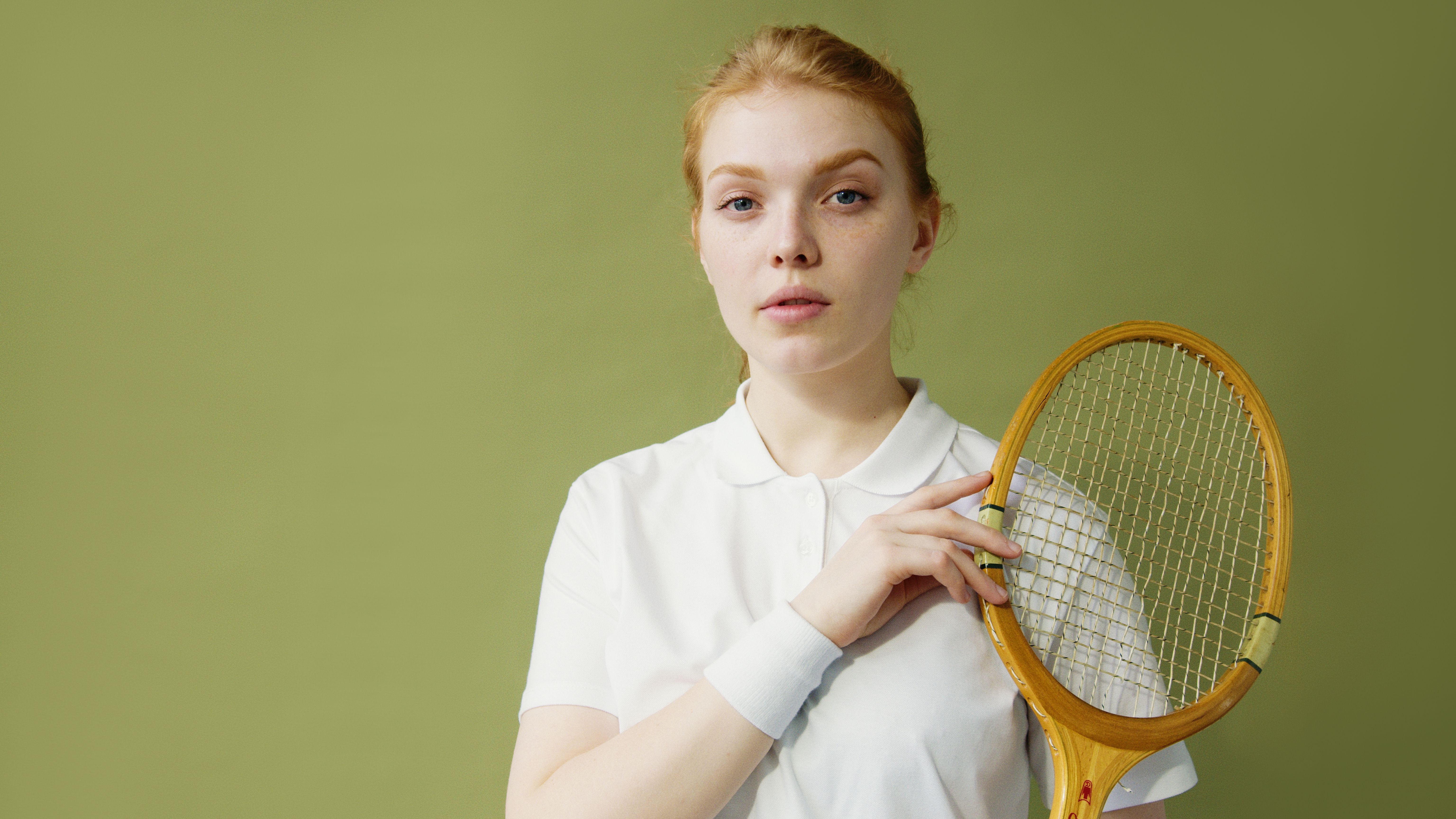 Young woman with red hair holding a tennis racket against green backdrop.