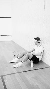Black and white photo of a man in activewear resting with a racket on a squash court.