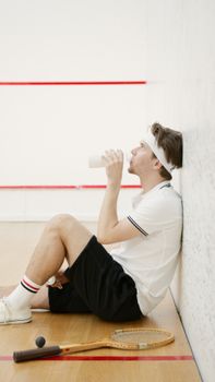 A young man takes a break and drinks water after playing squash indoors.