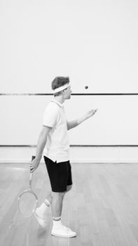 Athletic man in a squash court tossing a ball, captured in black and white.