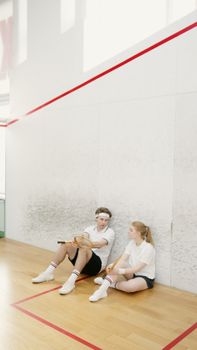 Two young adults taking a break on a squash court, holding rackets and looking relaxed.