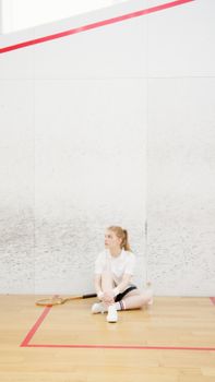 A woman in sportswear sits on a squash court floor with a racket.