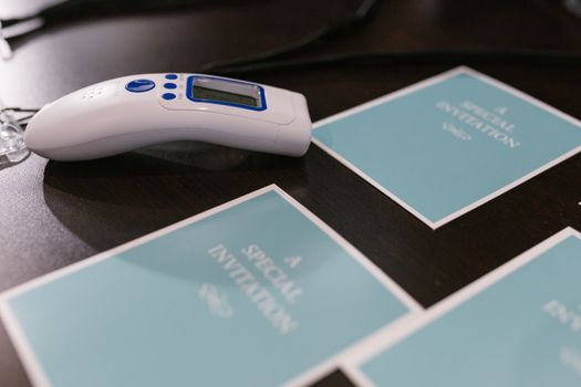 A close-up view of invitations and an infrared thermometer on a wooden surface, symbolizing event preparation and safety.