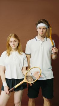 A young couple in sportswear poses seriously with tennis rackets against a brown background.