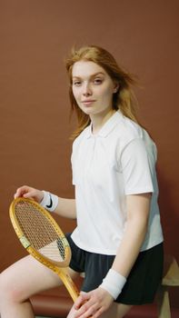 Woman in sportswear posing with tennis racket against brown backdrop, showcasing athletic style.