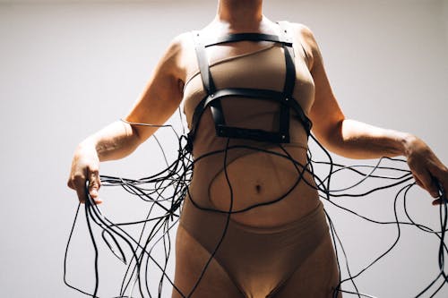 Crop of slim female in beige swimwear with bare shoulders holding long black wires standing straight on white background