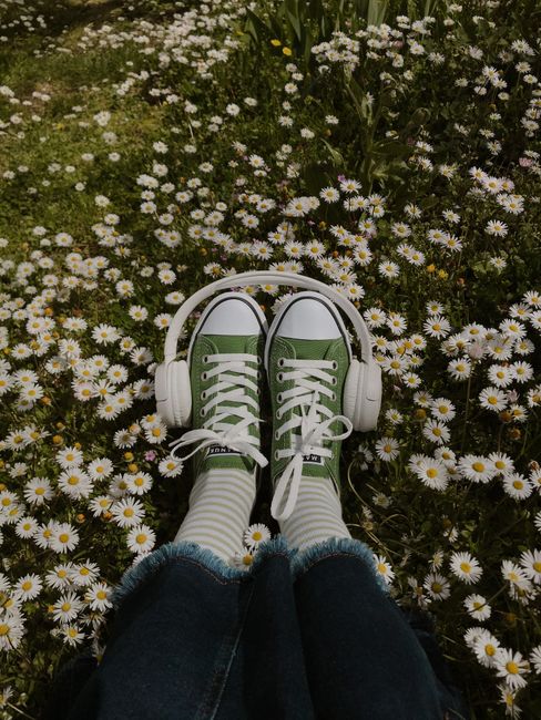 From above of crop anonymous female in jeans on glade with blossoming chamomiles with wireless headphones on legs