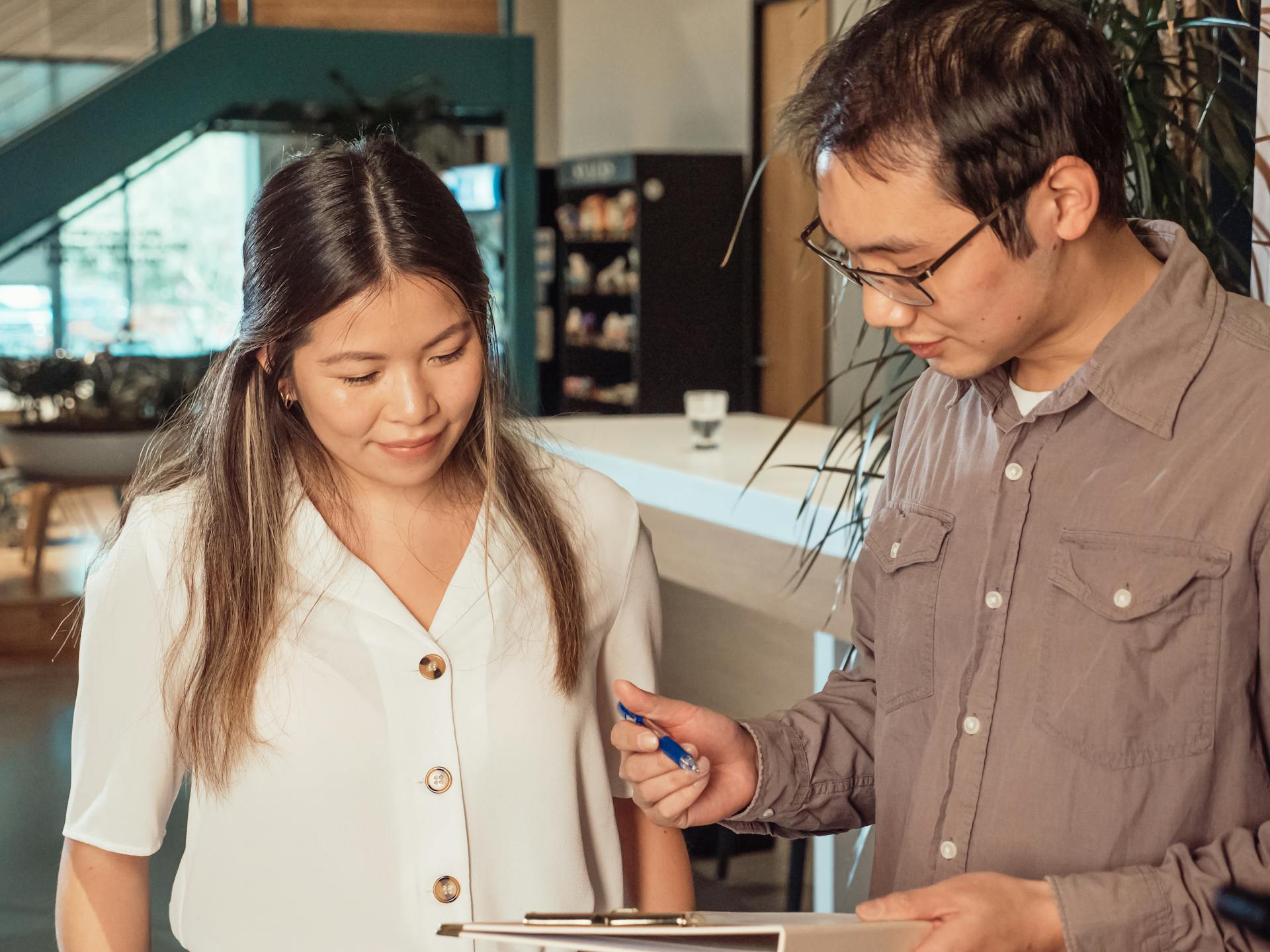 Asian man and woman in business attire talking and reviewing documents at the office. Bright indoor setting.