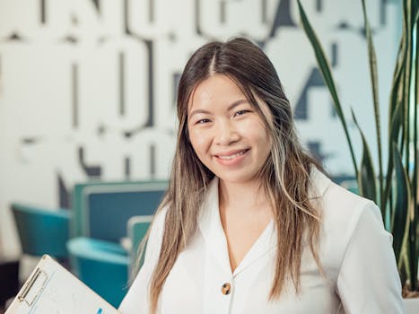 A cheerful woman in a white blazer holding a clipboard, captured indoors.
