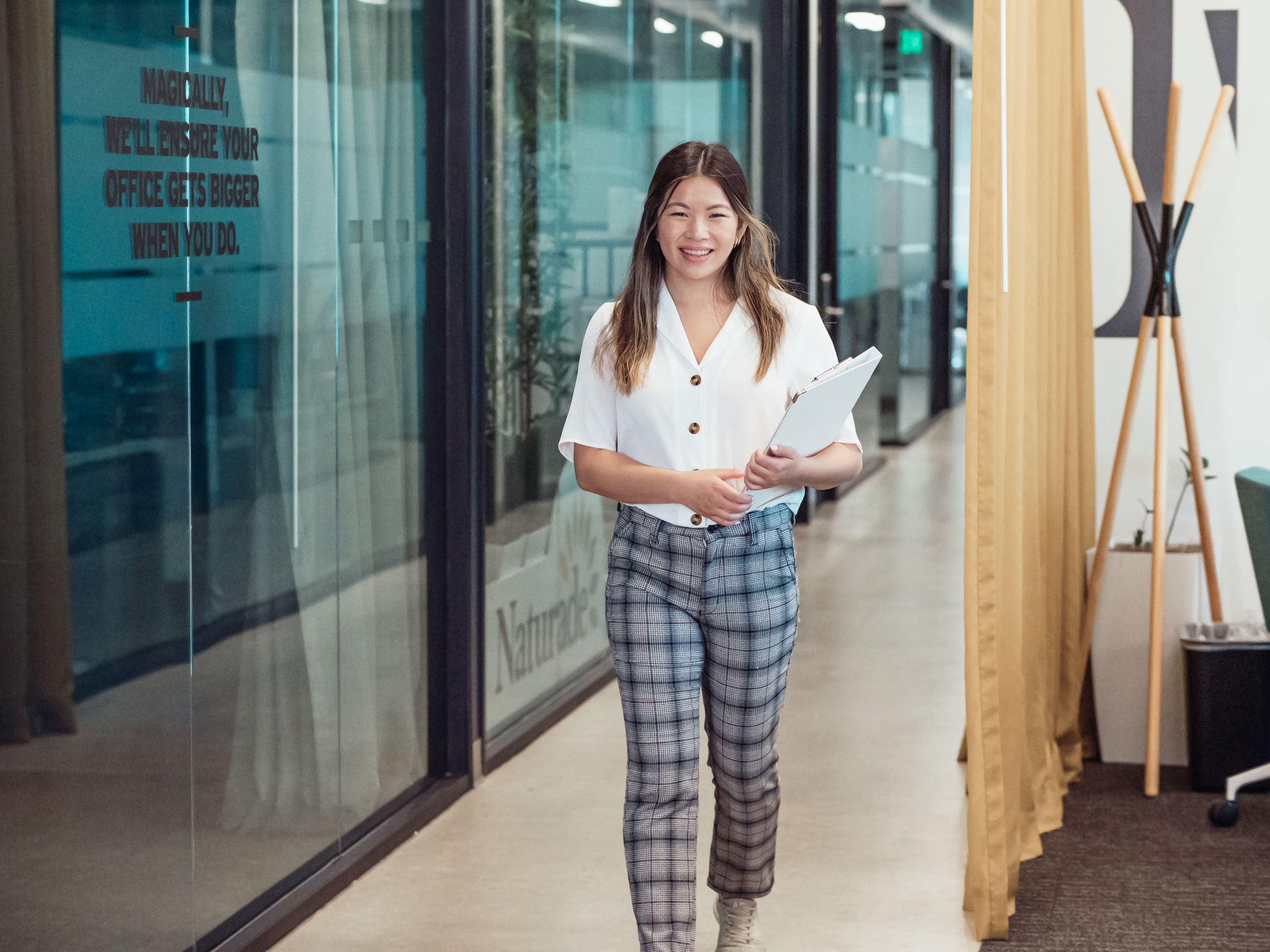 Confident Asian woman walking through a modern office hallway holding documents.