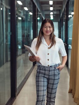 Smiling woman holding documents confidently walking in modern office corridor.