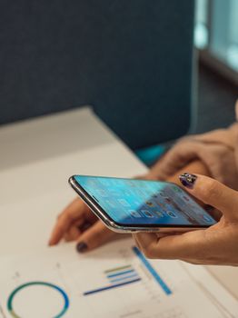 Close-up of hands holding a smartphone while analyzing financial graphs on paper.