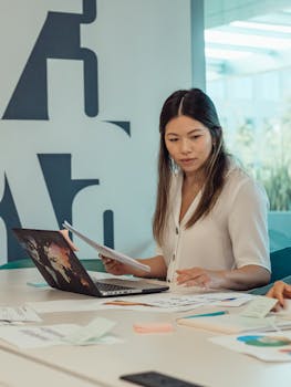 Asian woman in a modern office environment, reviewing paperwork with a laptop on the desk.